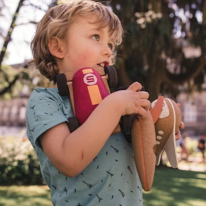 A blonde child is outside on a sunny day holding a collection of Elou cork toys in his arms
