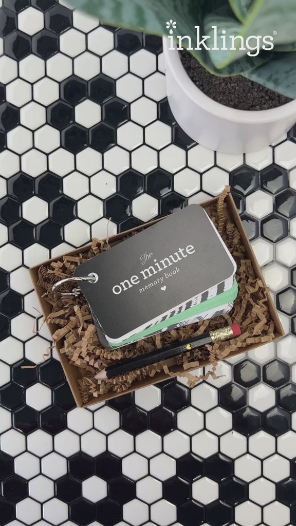 Video showing a light skinned person's hands taking the One Minute Memory Book out of the box and flipping through the pages to show them to the camera. The box is on a tiled surface with a black and white geometric design and there is a houseplant in a white pot in the corner.