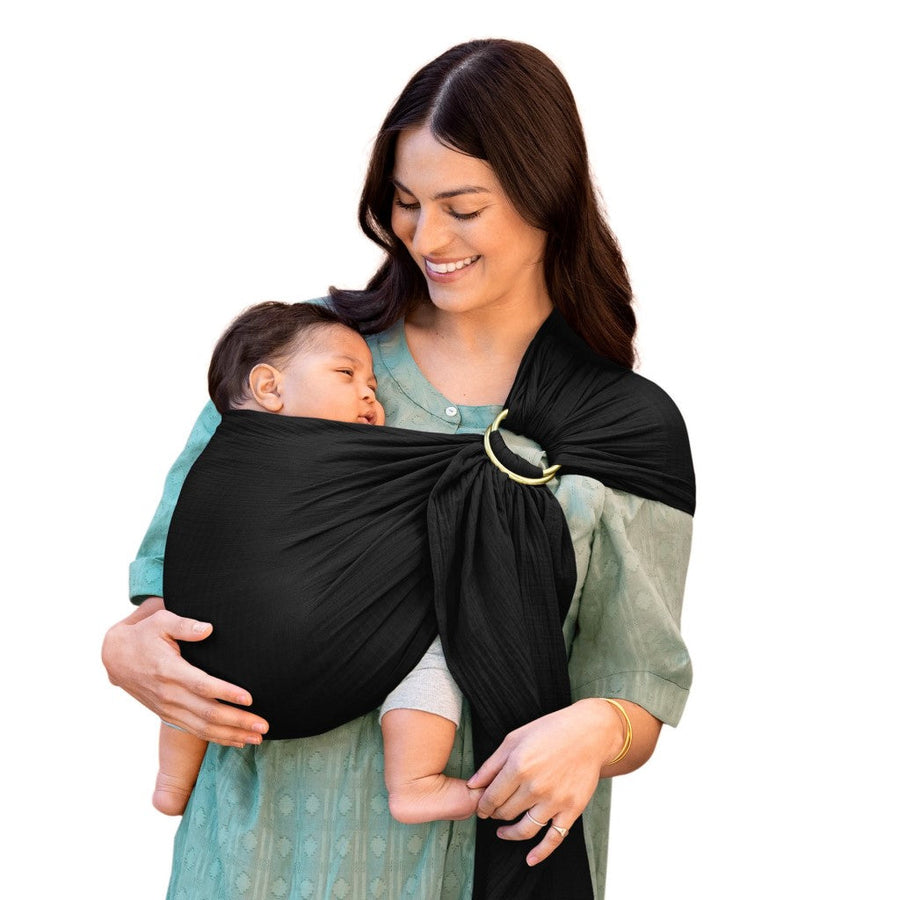 Picture of a woman holding a baby using a Moby Wrap Ring Sling Carrier in Onyx. She has long dark hair and a medium skin tone and is wearing a blue-green dress. She is smiling and looking down at a baby with short dark hair and a medium skin tone in a legs-out position in the black ring sling
