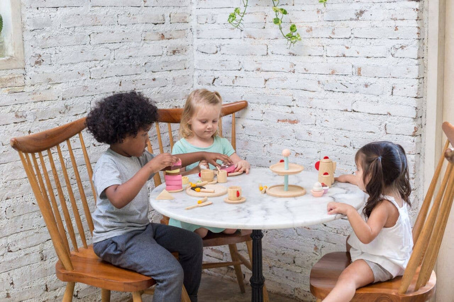 Three childrenhaving a tea party with wooden toys at a table against a white brick wall.