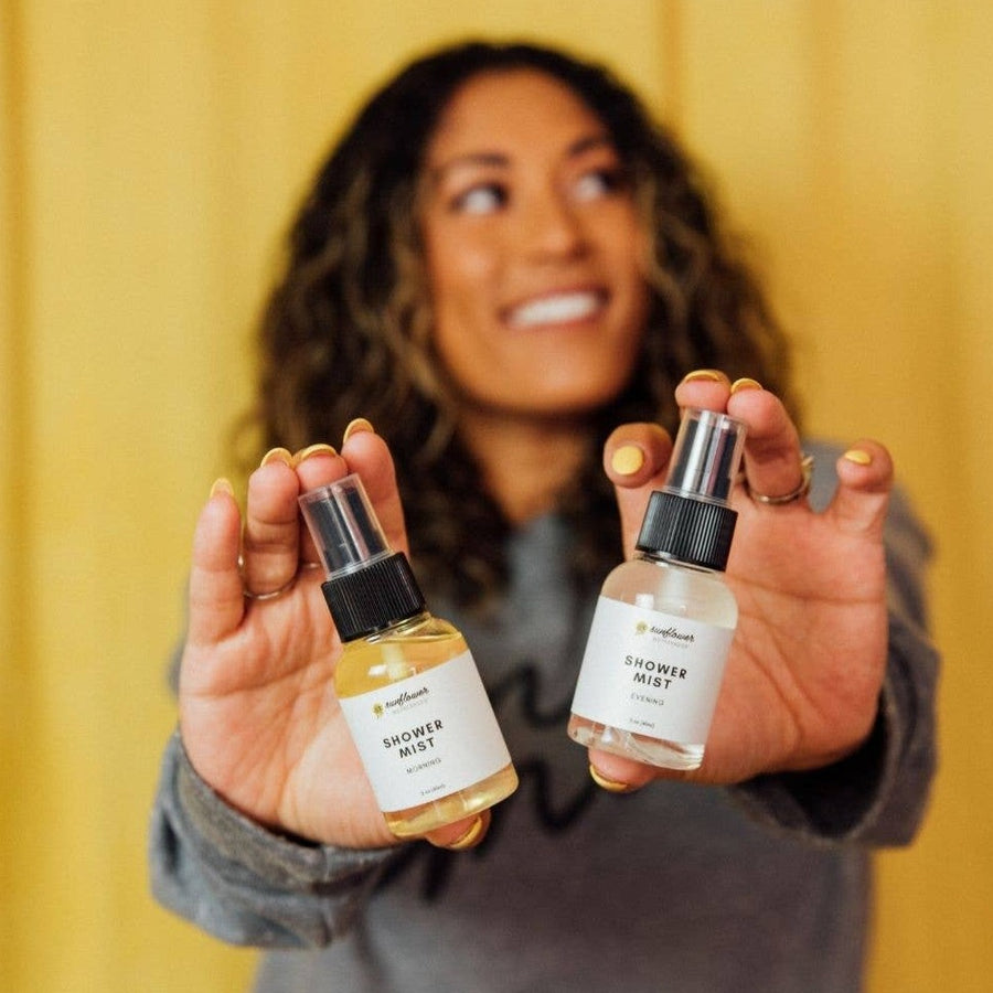 Image shows the shower mist bottles being held towards the camera one in each hand, by a person with long wavy dark brown hair, a medium skin tone, and long pale yellow painted fingernails. She is standing against a yellow backdrop looking up to the right and is wearing a light gray sweatshirt and rings on each ring finger