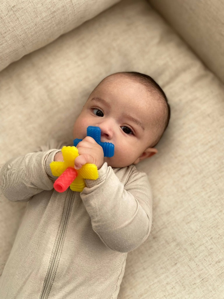 Picture of a baby with medium light skin and dark baby hair, holding the rainbow Teensy Tubes teether with one end in their mouth. They are wearing a beige zippered footie and lying on an upholstered beige surface