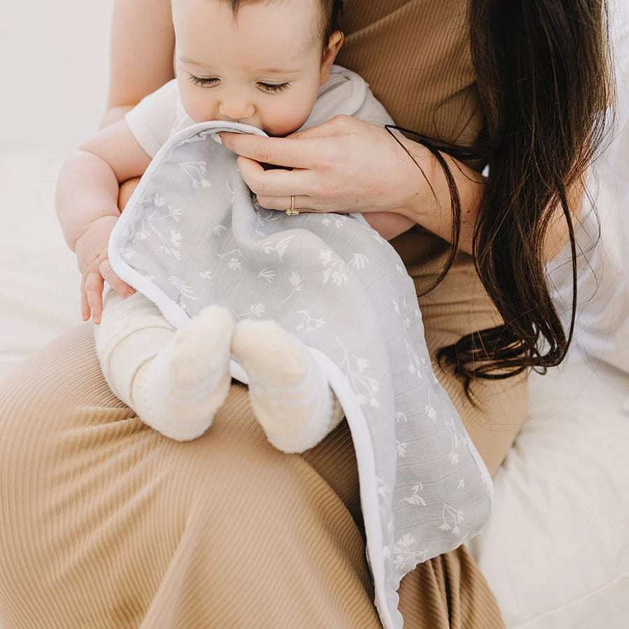 Picture of a baby sitting in a woman's lap, who is wiping their mouth with the Prairie burp cloth. Baby has a light skin tone and brown hair and is wearing a white short sleeved top with white pants and socks. The woman has a light skin tone and long brown hair and is wearing a gold ring and a ribbed light brown top and skirt.