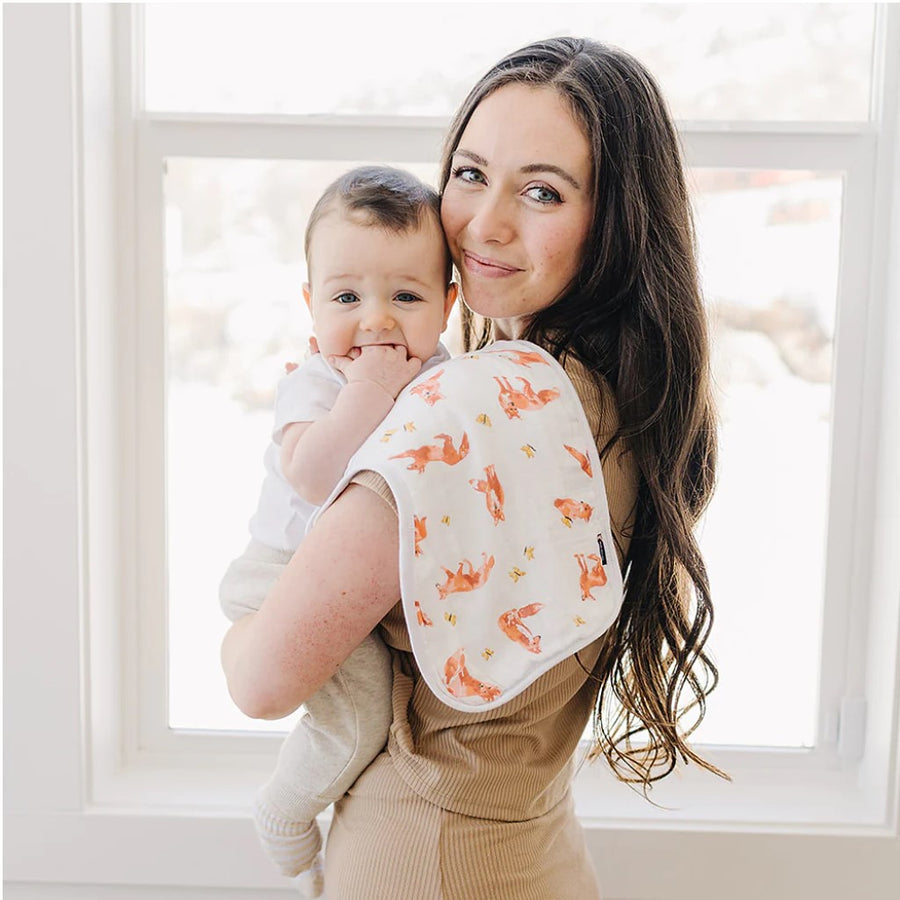 Picture of a woman standing in front of a window, holding a baby with the Fox Tales burp cloth draped over her shoulder. She has a light skin tone and long brown wavy hair, and is wearing a beige colored short sleeved top and bottoms and smiling over her shoulder at the camera. The baby has a light skin tone and short brown hair and is looking at the camera with the fingers of one hand stuck in their mouth. Baby is wearing a short sleeved white top with cream colored pants and socks.