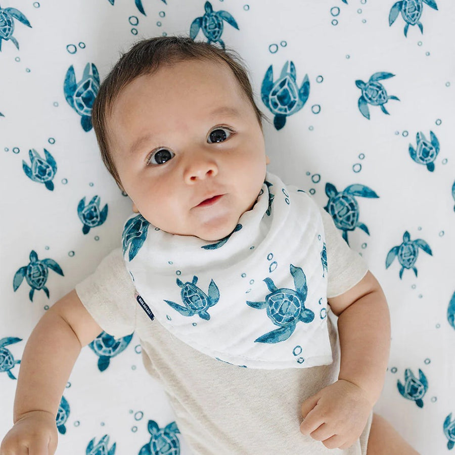 Picture of a fair skinned baby with large eyes and brown hair, looking at the camera while lying on their back on a Sea Turtles sheet, wearing a Sea Turtles bib over a short sleeved white bodysuit