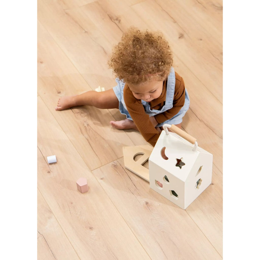 Picture showing a toddler sitting on a wood floor, reaching inside the shape sorter house. The child has a medium skin tone and reddish brown curly natural hair, a long-sleeved brown top, and denim overalls