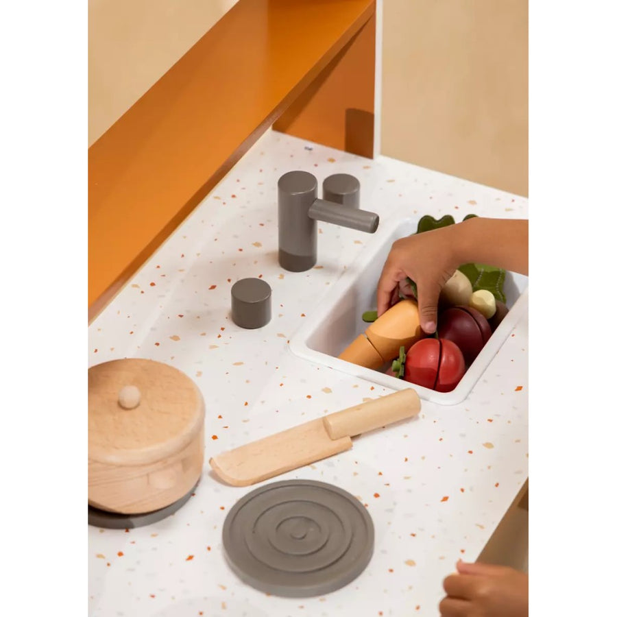 Picture shows a play kitchen with a white flecked counter and a wooden pot on a burner. The wooden veggies are in the sink with a child's hand reaching in and picking up the carrot