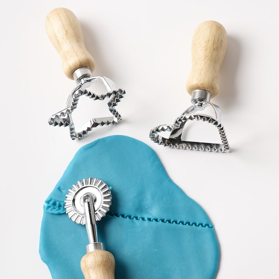 Picture of some flattened blue play dough on a white surface, with a textured line across it from the cutting wheel tool, which is lying on top of the dough. The star cutter and heart cutter are on the surface above the dough.