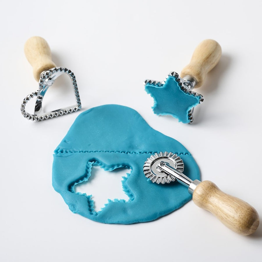 Picture of some flattened blue play dough on a white surface, surrounded by the Eco-kids Ecodough Cookie Cutter Tools. The dough has a star shape cut out from it from the star cookie cutter. The star cookie cutter still has blue dough in the star shape filling up the cutter. There is also a textured line across the dough from the cutting wheel tool.
