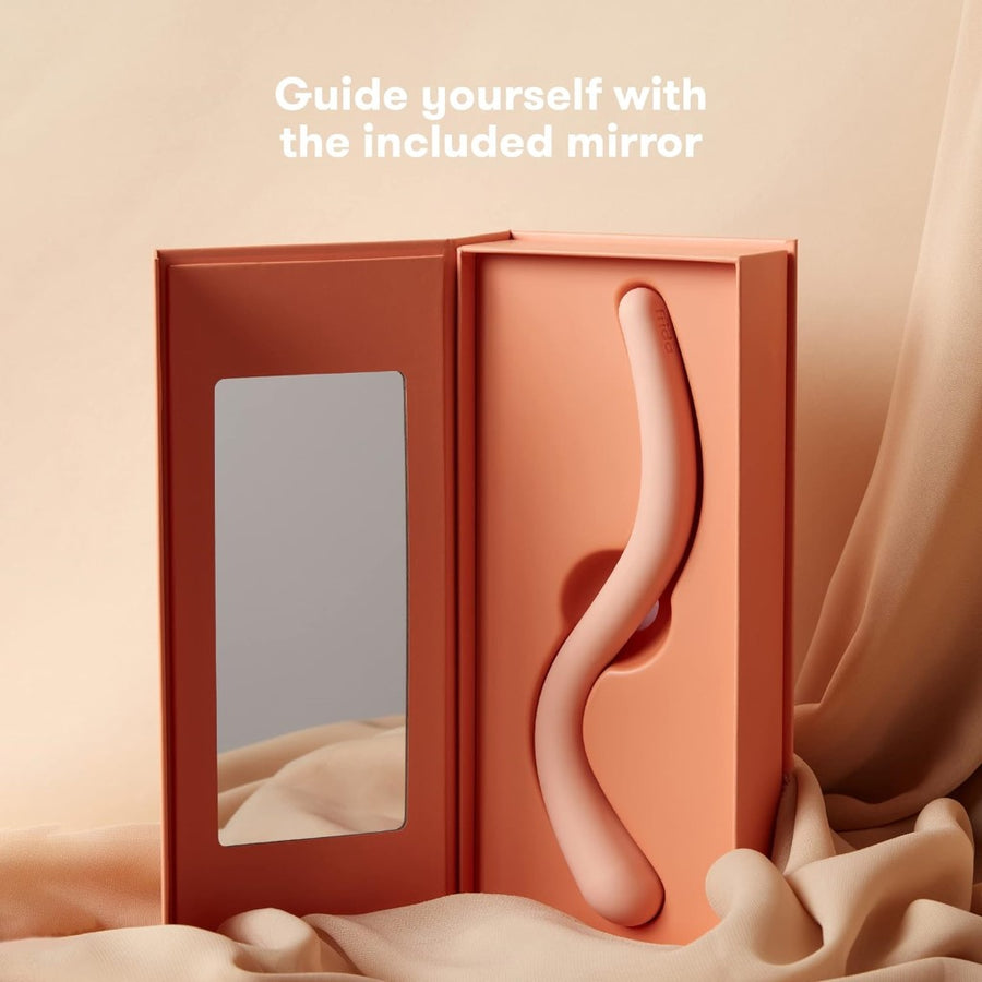 Image shows the open box containing the perineal massage wand with the mirror inside the lid, against a pale pink backgroun and sitting on light pink draped fabric. Text at the top reads 