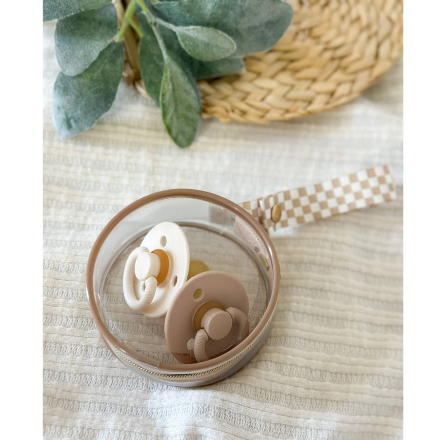 one cream colored and one rose colored pacifier inside the itzy paci case, on a white tablecloth with a wicker basket and a leafy sprig of a plant in the upper part of the photo