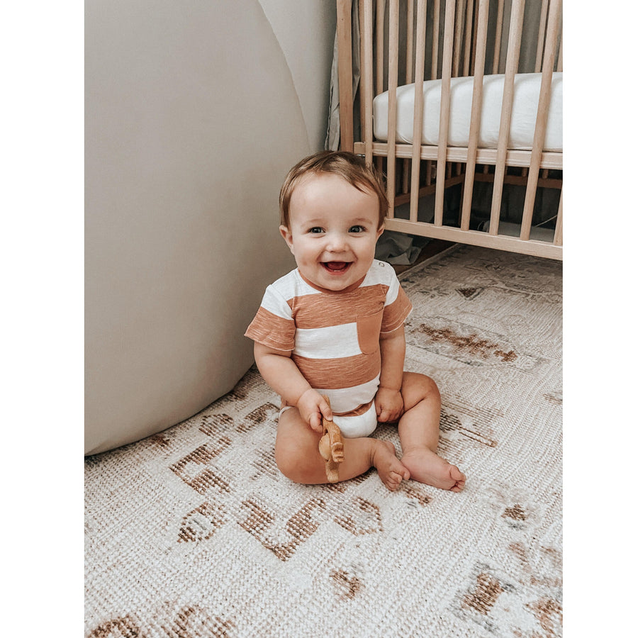 Picture of a baby wearing the L'ovedbaby Slub Jersey Crewneck Bodysuit in Adobe Stripe. The baby has a light skin tone and brown hair and is laughing while looking at the camera, seated in butterfly pose on a faded patterned carpet next to a wooden crib and in front of a gray wall against which a large round gray padded object is resting, perhaps a play mat or cushion. The baby is holding a small wooden toy.