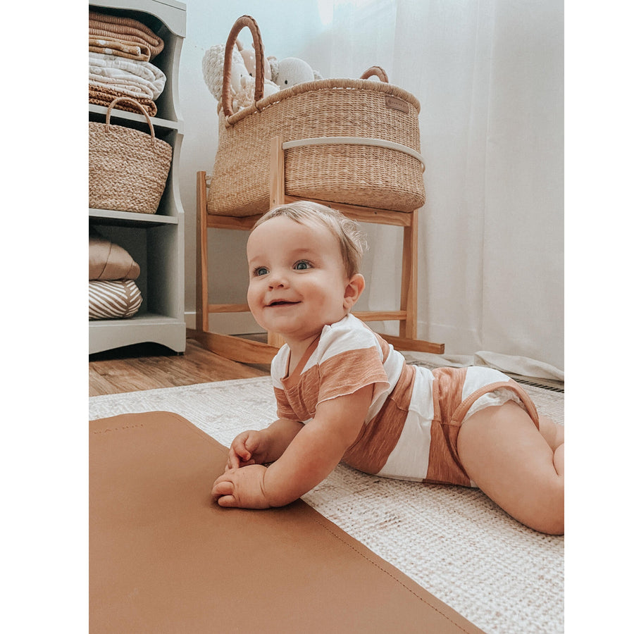 Picture of a baby wearing the L'ovedbaby Slub Jersey Crewneck Bodysuit in Adobe Stripe. The baby has a light skin tone and short light brown hair, and is on their stomach raising themselves up with their hands and looking to the left. They are on a beige carpet over a hardwood floor at the edge of an adobe-colored play mat, in front of a wicker bassinet on a wooden bassinet stand and a wood shelf with folded linens.