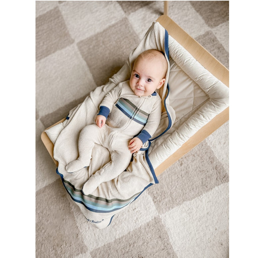 Picture of a baby with light skin and dark eyes, looking at the camera while wearing the footie and laying on a matching blanket in a wood rocker board with a cream colored pillow. The rocker sits on a checkered carpet of cream and stone gray.