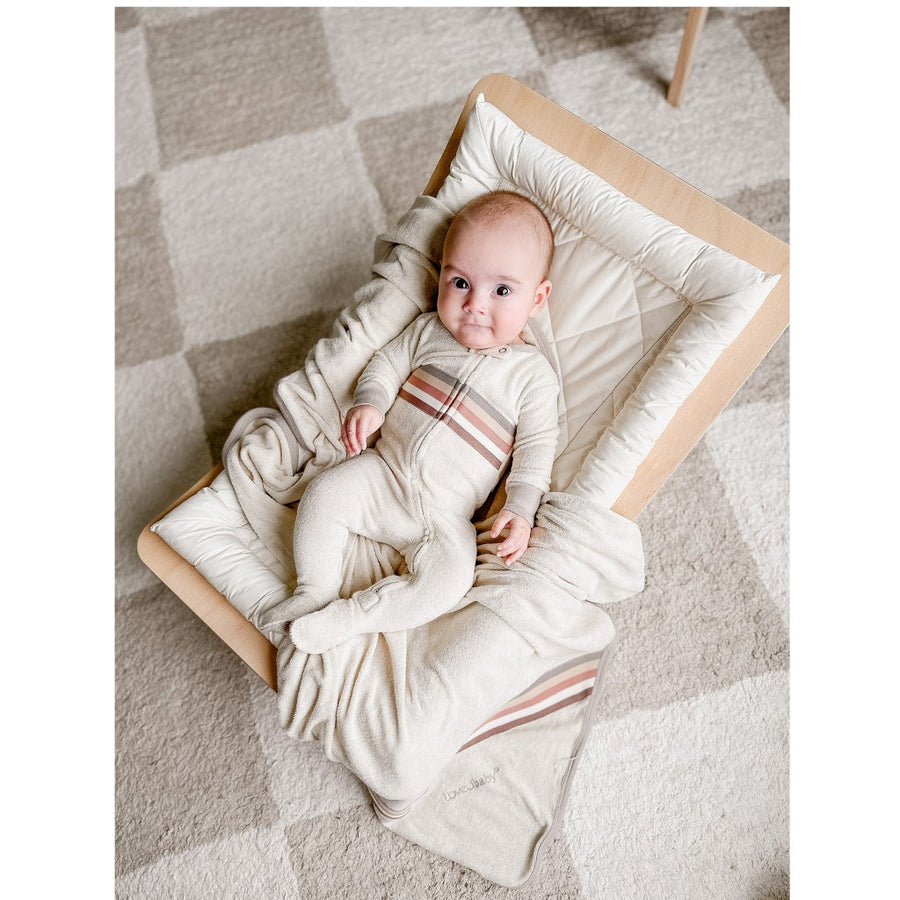 Picture of a baby with light skin and dark eyes, looking at the camera while wearing the footie and laying in a wooden rocker with a cream colored pillow and matching blanket. The rocker is sitting on a checkered carpet that is cream and stone gray.