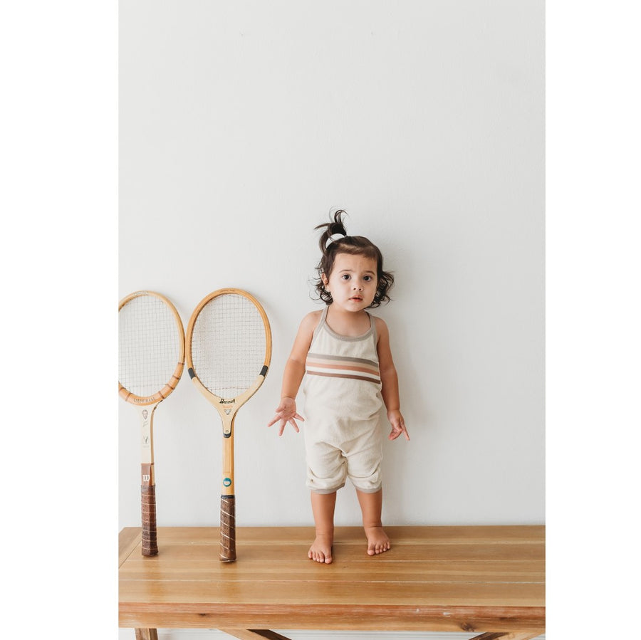Picture of a toddler wearing the overalls, looking at the camera while standing on a natural finish wood table against a gray wall.  The child has a light skin tone and dark eyes and hair with a half-up ponytail sticking up. There are two tennis rackets leaning against the wall next to the child.