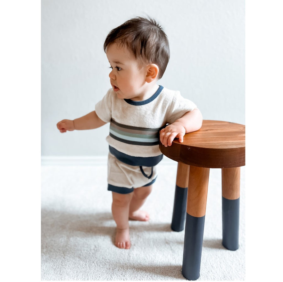Picture of a toddler with light skin and short dark hair, wearing the shorts and tee set while standing on a white carpet and leaning with one arm on a wooden stool and looking to the left.