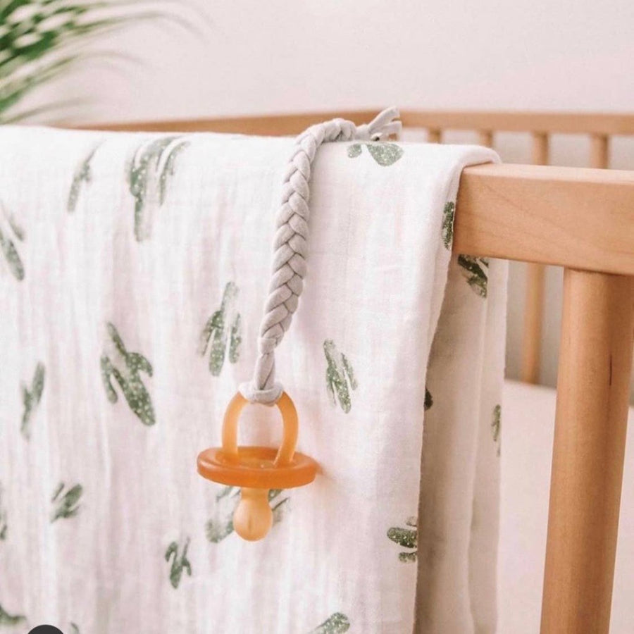 Image of a ecopacifier attached to a braided white cord pacifier clip, hanging over the edge of a wooden crib with a saguaro print swaddle over the rail