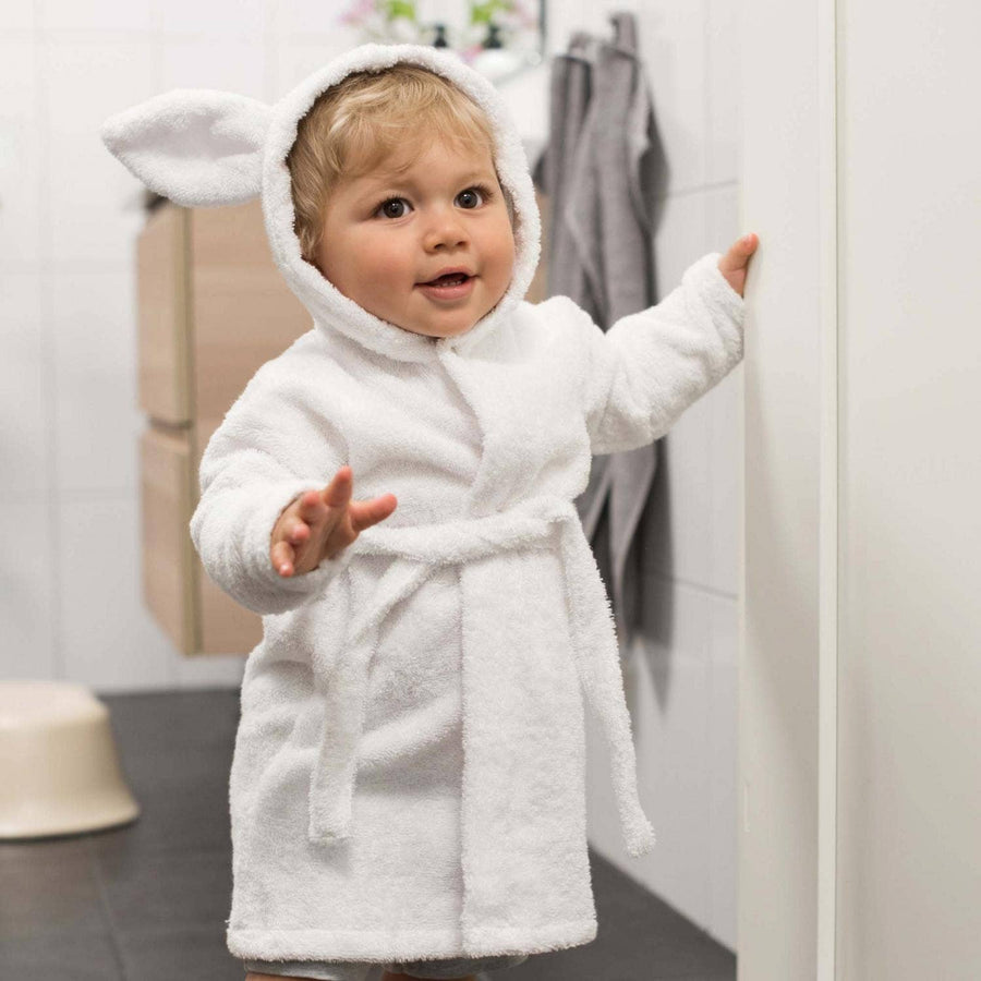Baby in a white bathrobe with animal ears standing in a bathroom.