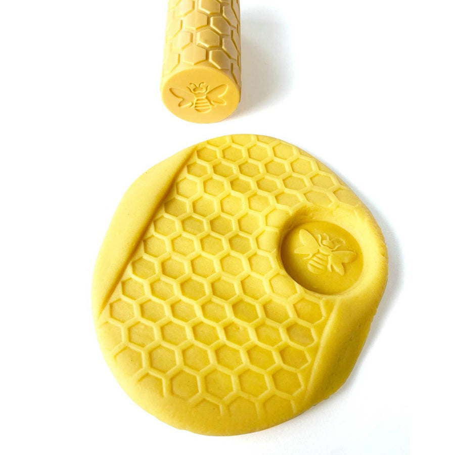 picture of yellow play dough that has been embossed with a honeycomb pattern and an impression of a honeybee, with the Bee silicone roller above it on a white background
