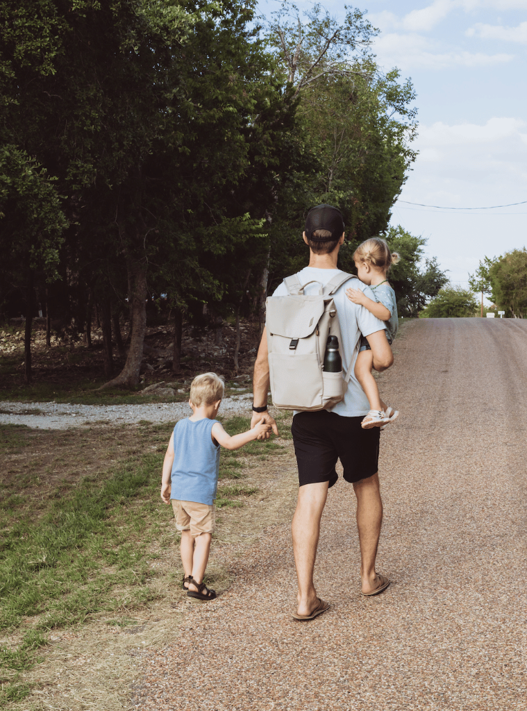 Picture from the back of a light-skinned man with short brown hair wearing the taupe backpack and walking down a gravel road lined with trees. He is carrying a small light-skinned blonde haired child with pigtail buns and leading by the hand another child with light skin, blonde hair, a blue tank top and beige shorts. The man is wearing a black baseball cap, a black watch, a light gray t shirt, black shorts, and brown sandals.