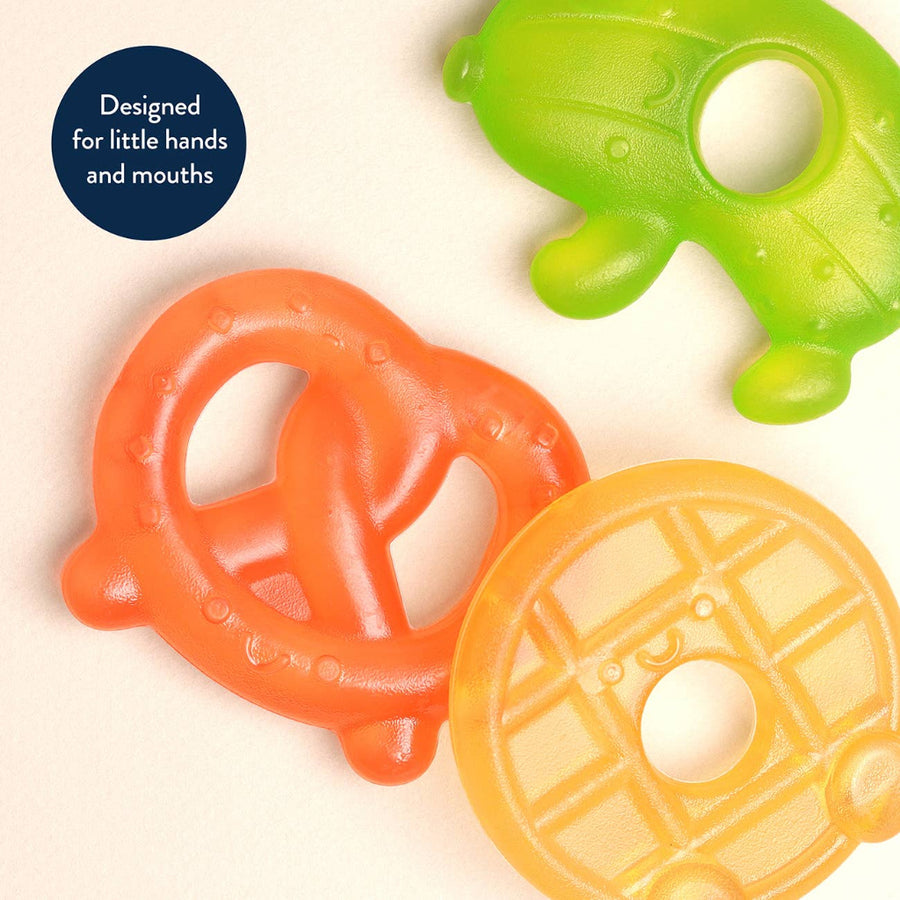 Three colorful silicone teethers shaped like a pretzel, waffle, and pickle on a beige background.