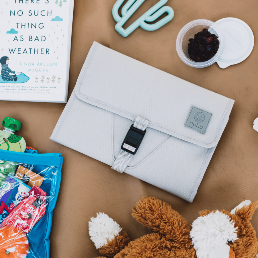 Picture of the taupe changing wallet on a brown leather or faux leather surface with other items including a mint cactus shaped teether, a book, a snack cup of raisins, a stuffed fox toy, and the backpack's zipper pocket bag containing snacks