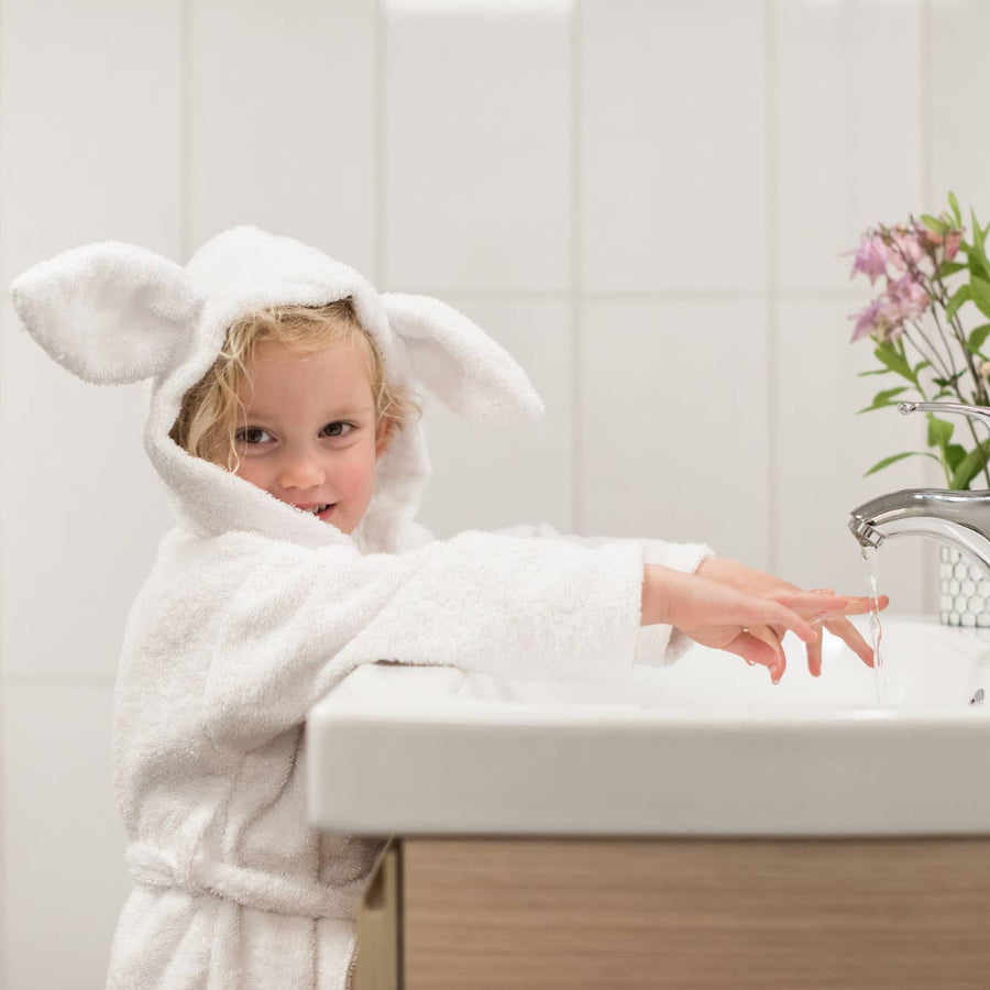 Child in a white bathrobe with bunny ears washing hands in a sink.