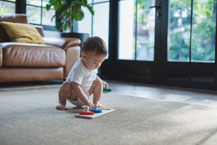 Baby playing with colorful toys on a carpeted floor in a living room.