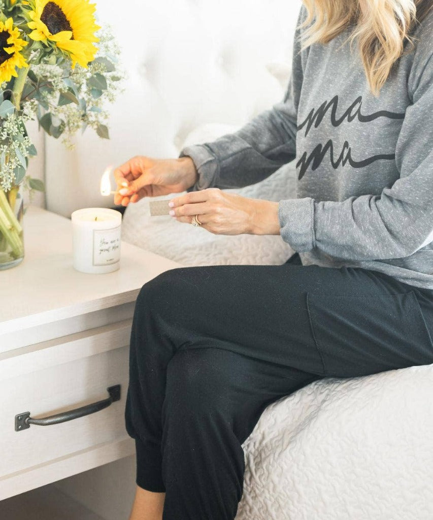 Image shows a light-skinned person with long blonde hair, wearing black pants and a gray Mama sweatshirt, seated on a bed with a white comforter while lighting the Great Mom candle. The candle is on top of a white bedside cabinet with a bouquet of sunflowers and eucalyptus in a clear glass vase
