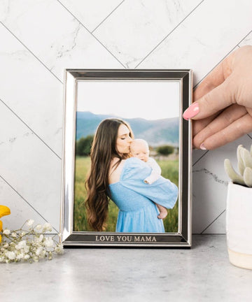 Picture of a light-skinned hand with light pink fingernails, setting the photo frame on a white table against a white tiled wall, with a sprig of white baby's breath and a small succulent in a white pot visible on the table. The frame has a photo of a woman with a light skin tone and very long brown hair, dressed in a light blue dress and holding a baby with a light skin tone and kissing it on the head. They are standing outdoors with a mountain backdrop