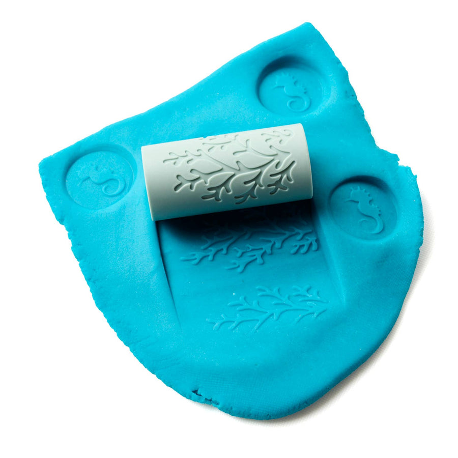 picture of some blue play dough that has been stamped in 3 places with a seahorse impression, with the Ocean silicone roller on top of it and showing the kelp impression in the dough from the roller