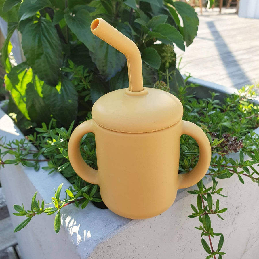 Yellow sippy cup with a straw on a plant-filled windowsill