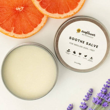 Image shows the salve tin open with the lid next to it, on a white surface with slices of pink grapefruit and sprigs of lavender flowers