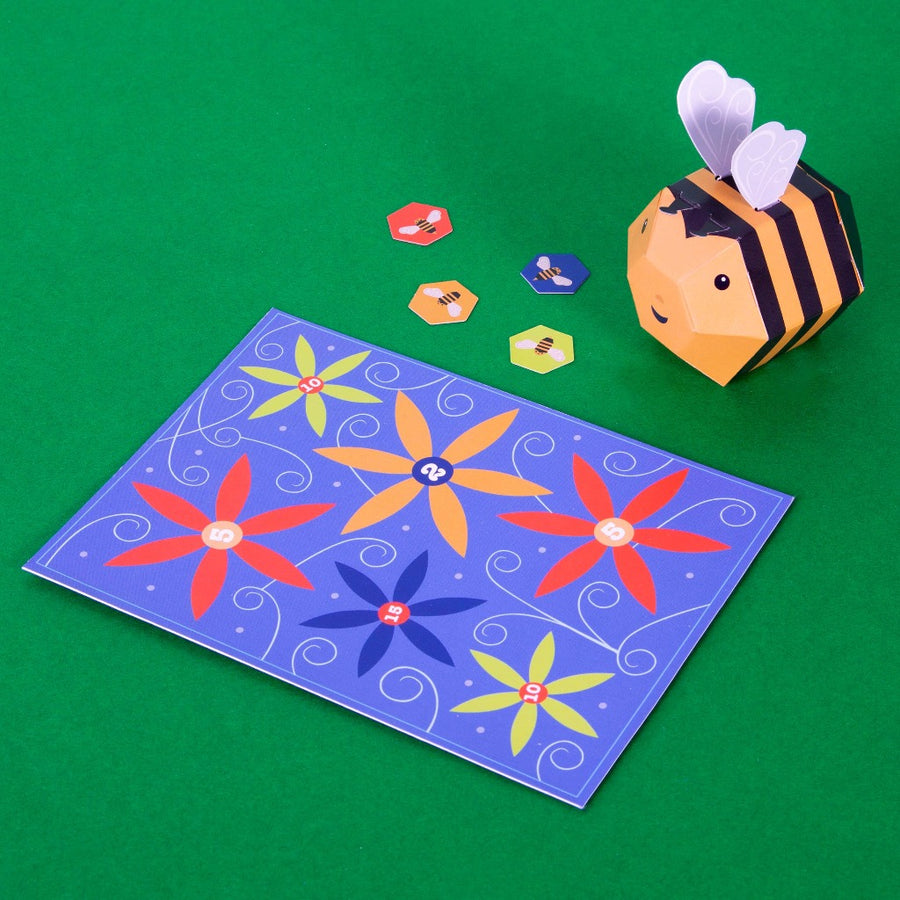 Image shows the pollination game board, several bee game pieces, and the assembled buzzy bumble bee, sitting on a green surface. The game board is purple with several large flowers 