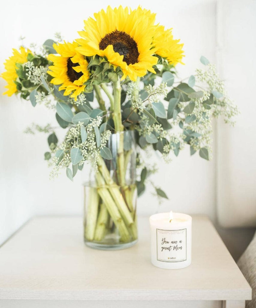 Image shows the Great Mom candle on a white table next to a large bouquet of sunflowers and eucalyptus in a clear glass vase