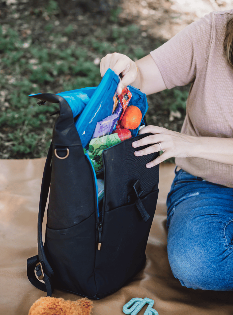 Picture of a light skinned person in blue jeans and a beige t shirt seated on a brown leather or faux leather surface outdoors, holding the open backpack next to them and removing one of the zipper pockets which is full of fruit and packaged snack