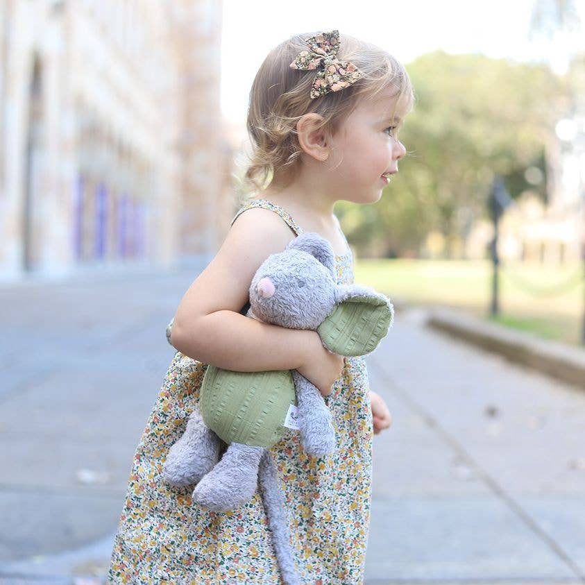 Picture of a girl with fair skin and short dirty blonde hair, wearing a sleeveless floral print dress with a matching hair bow, walking outdoors in profile from left to right, with the Tikiri Toys Organic Baby Mouse plush toy under her arm