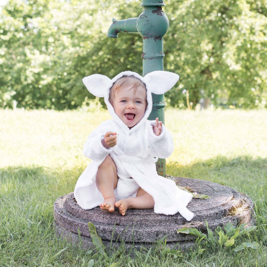 Baby in a white robe with animal ears sitting on a stone in a grassy area.