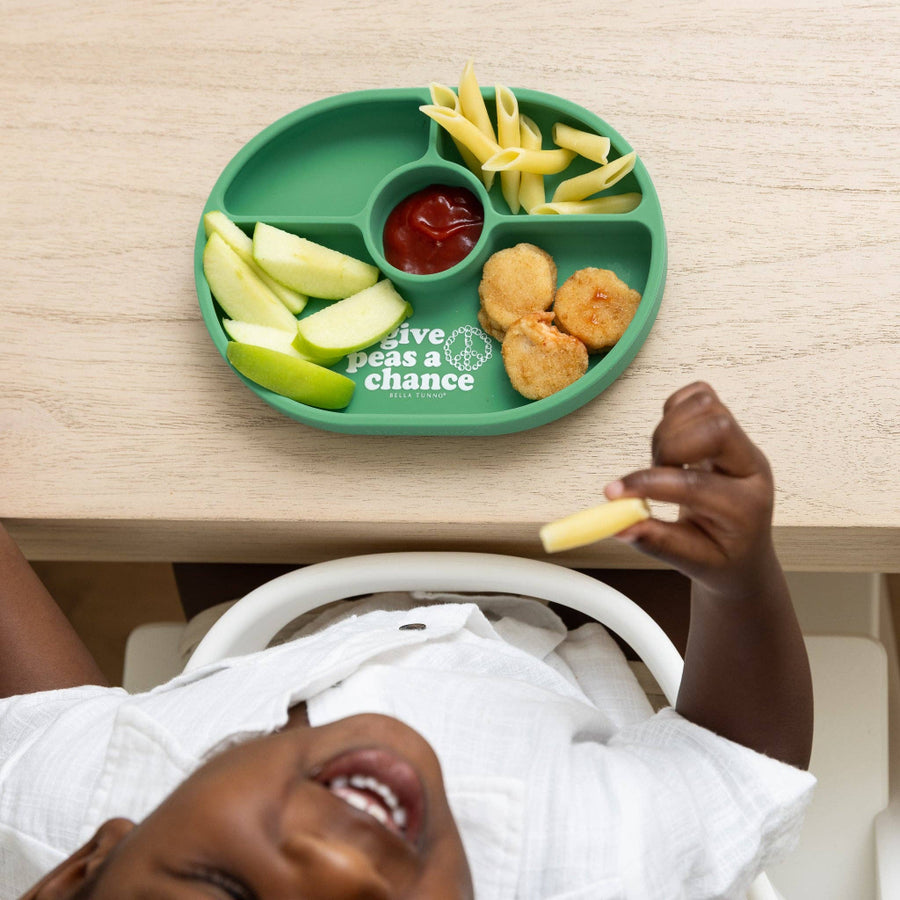 Child sitting at a table with a green plate containing food, including chicken nuggets, apples, and french fries.