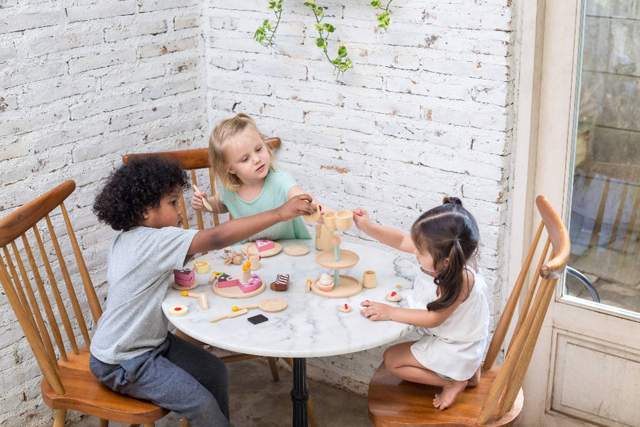 Three children playing with wooden toys at a table against a white brick wall.