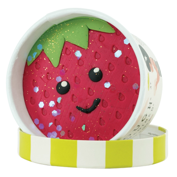Picture of the dough cup with the lid off. The surface of the dough is red like a strawberry with little impressions in the shape of seeds, a happy face, and a green top, with large and small glitter.