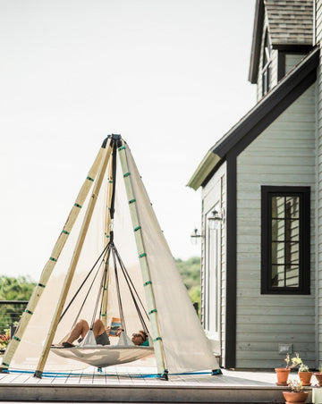 Person relaxing in a tepee-style hammock on a deck with a house in the background