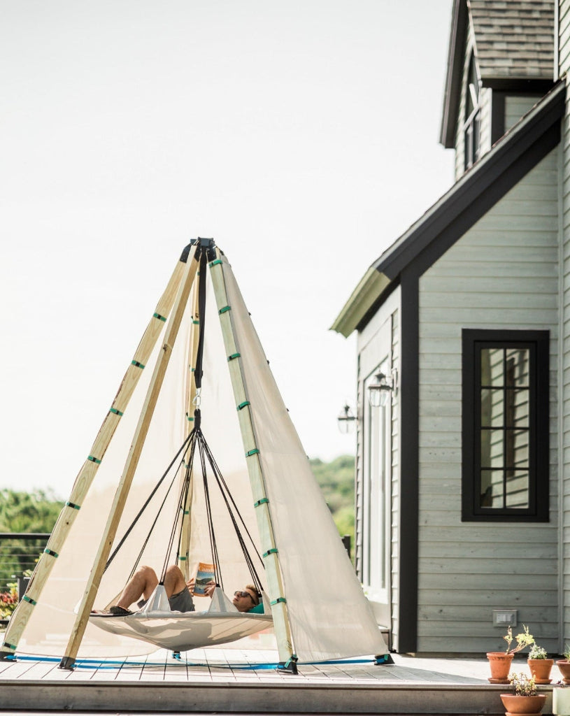 Person relaxing in a tepee-style hammock on a deck with a house in the background