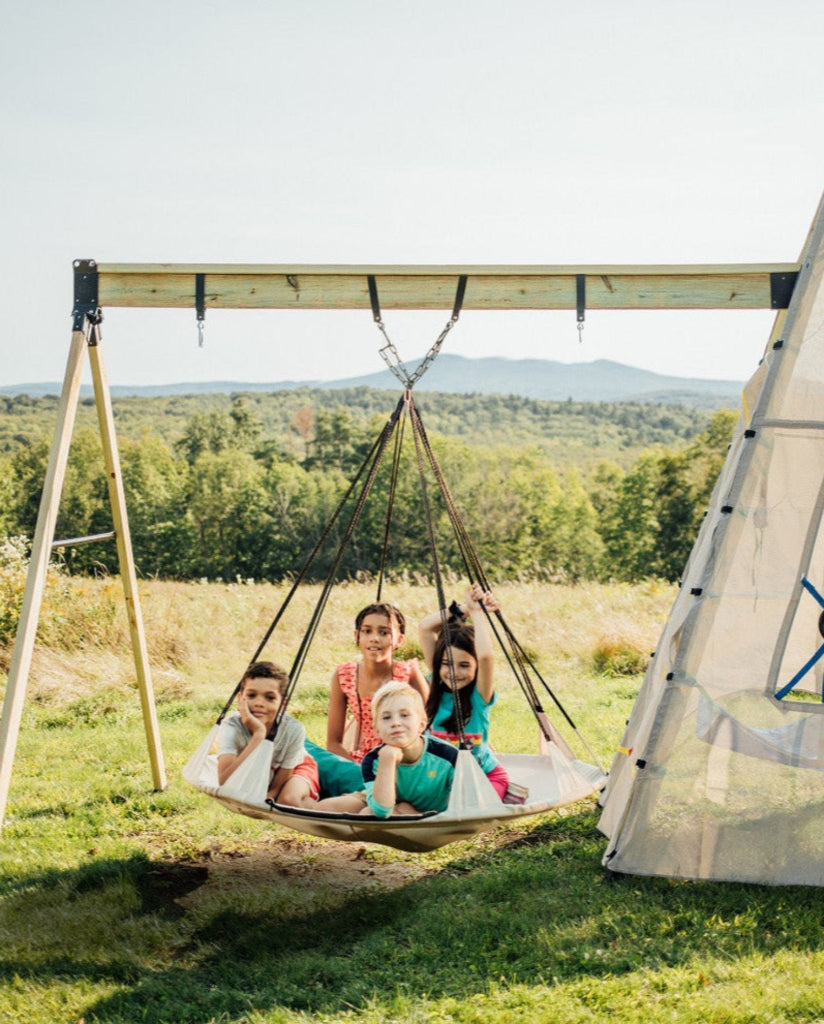 Children sitting in a hammock outdoors with a tent and scenic background