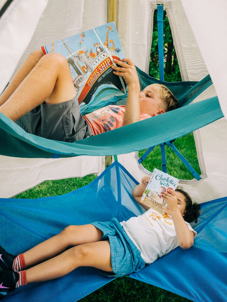 Two children reading books in blue hammocks inside a tent.