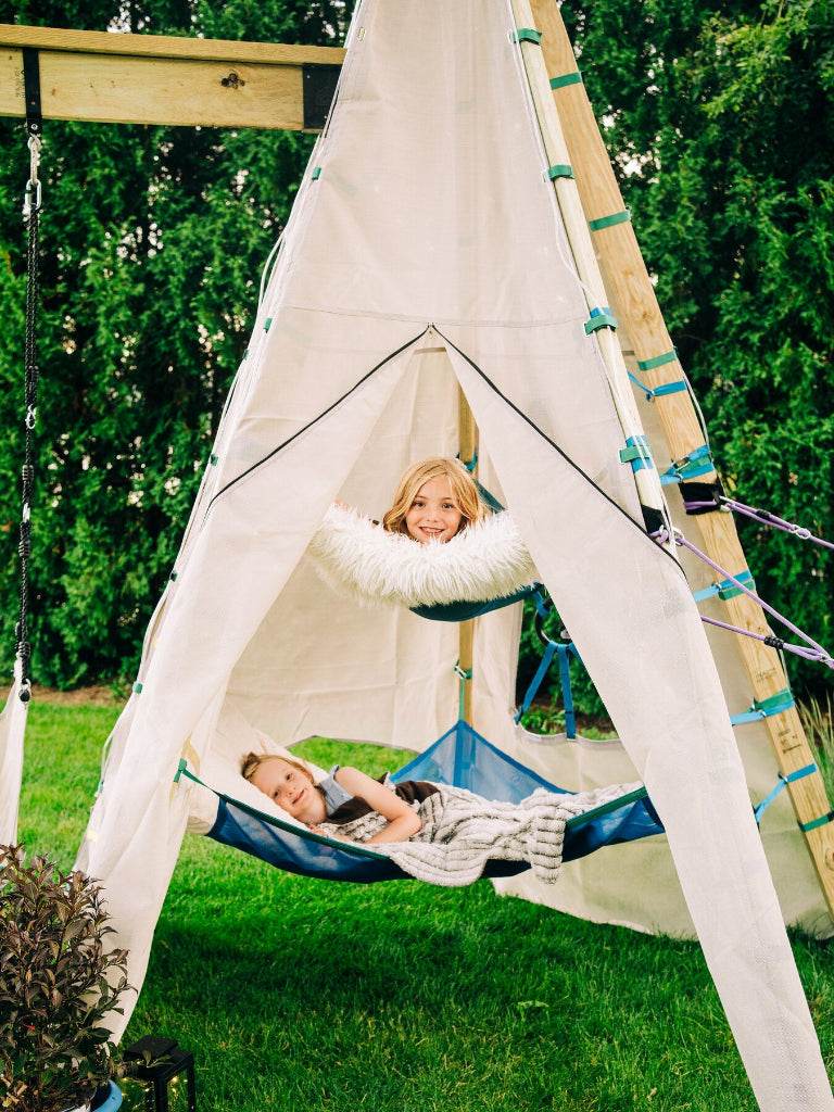 Children playing in a teepee with a hammock inside, surrounded by greenery.