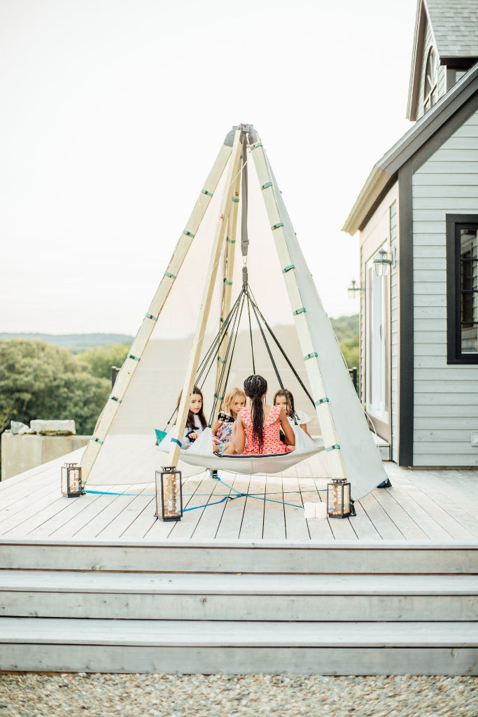 Children sitting in a teepee-style swing on a deck with lanterns around