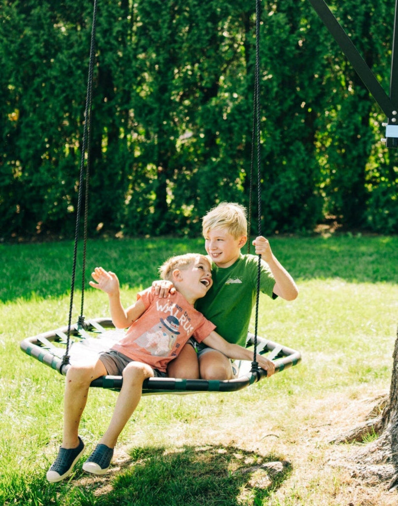 Two children on a swing set in a park with green grass and trees.