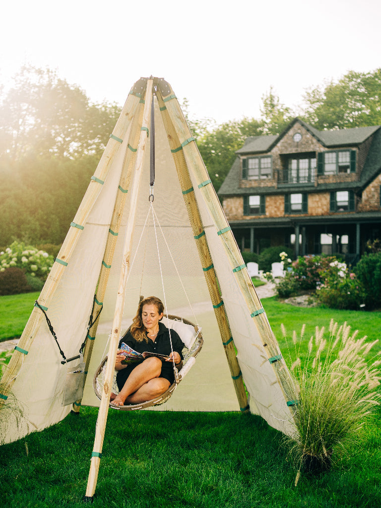 Person sitting in a hammock attached to a teepee structure in a garden setting.